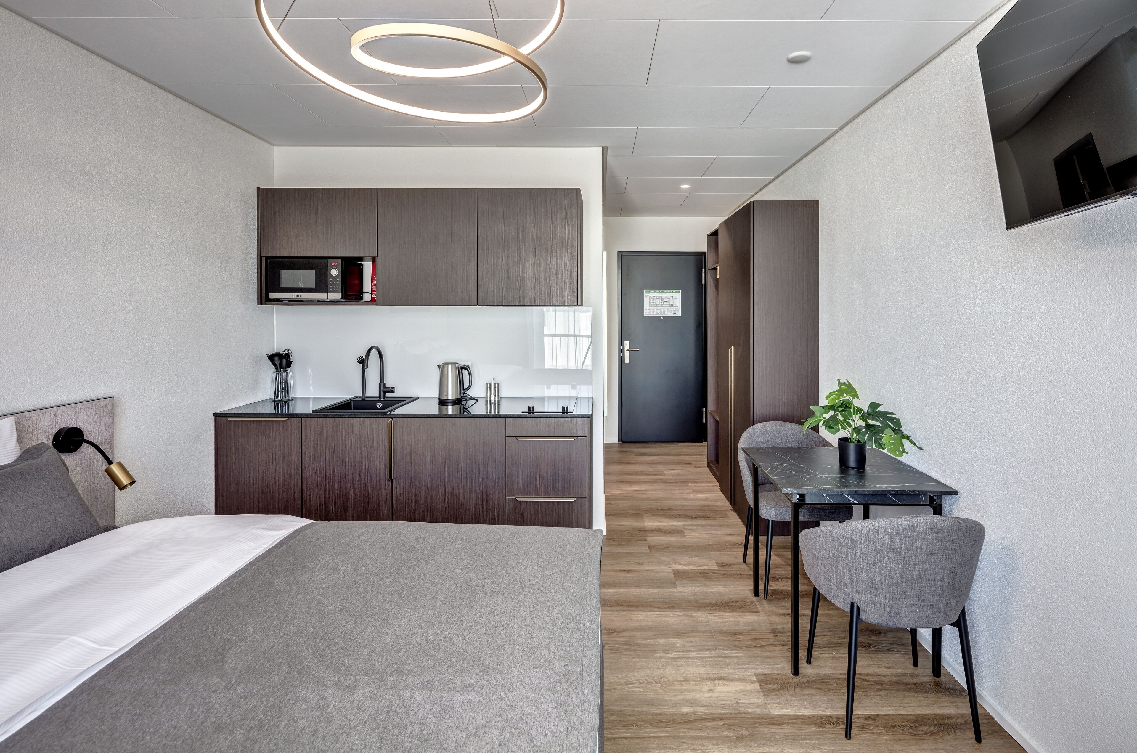A modern hotel room with a bed, a desk, a dining table, and a kitchenette.