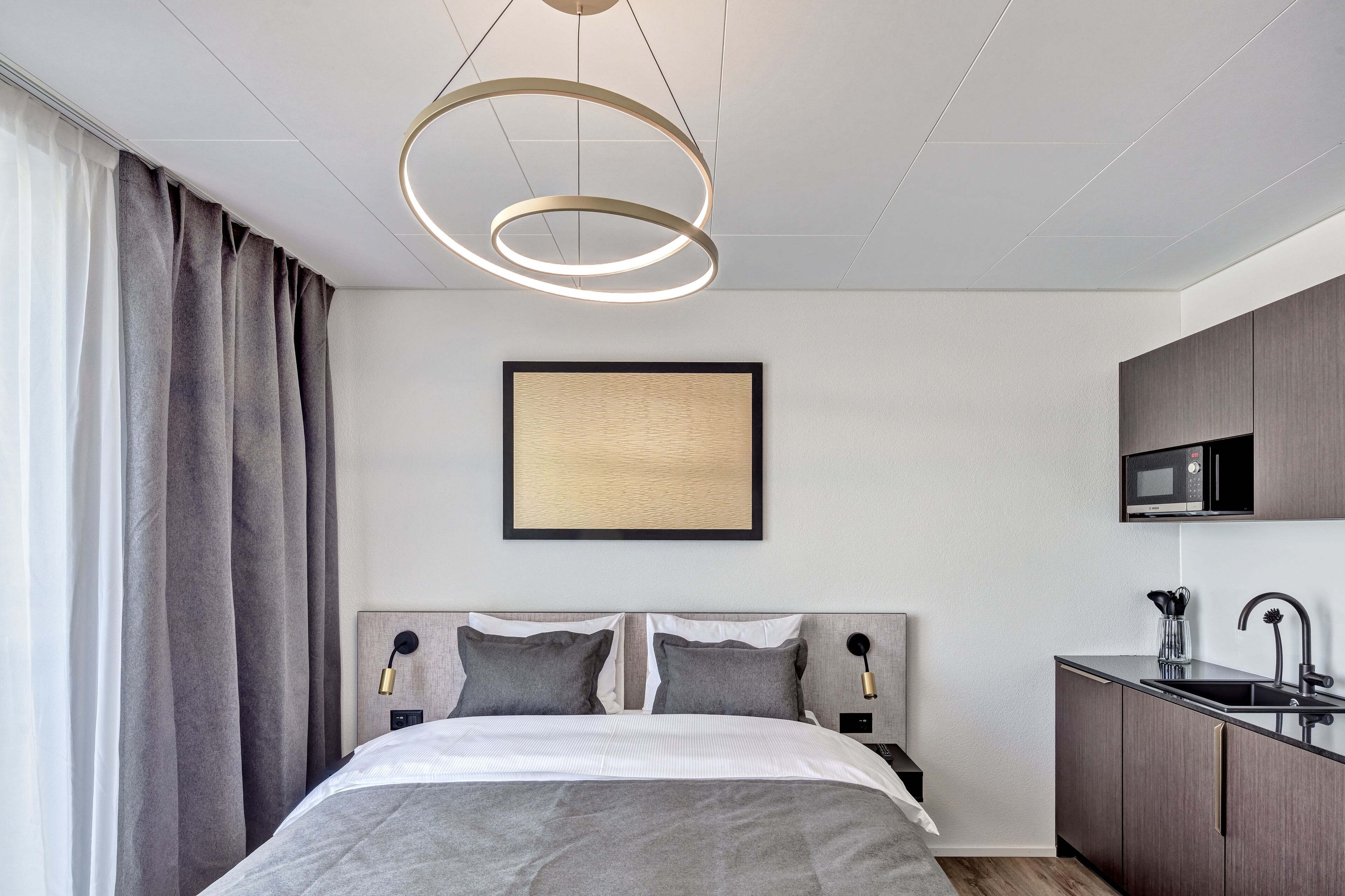 Quarto de hotel moderno com cama, mesa de cabeceira, televisão fixada na parede e área de cozinha compacta.