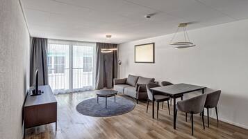 A modern living room with a sofa, dining table, and chairs, a TV, and a large window with curtains.