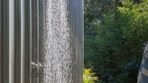 Una ducha con agua corriendo por una pared metálica vertical, un camino de hormigón y un jardín con diversas plantas.
