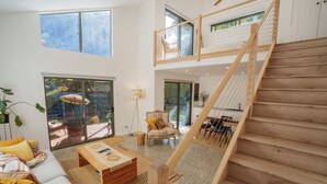 Amplia sala de estar con escalera de madera, ventanas grandes y vistas al exterior.