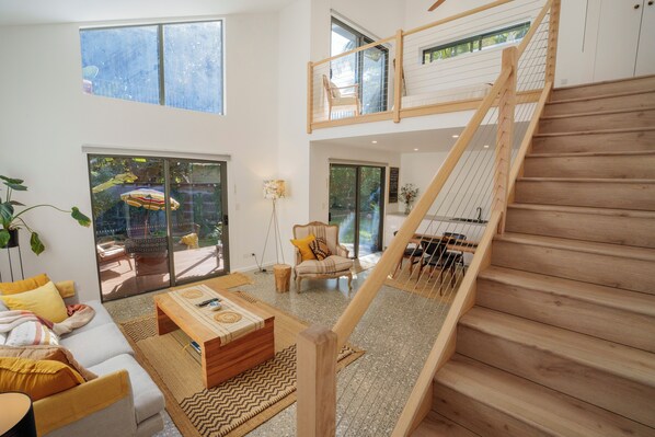 A spacious living area with a wooden staircase, large windows, and a view of the outdoors.