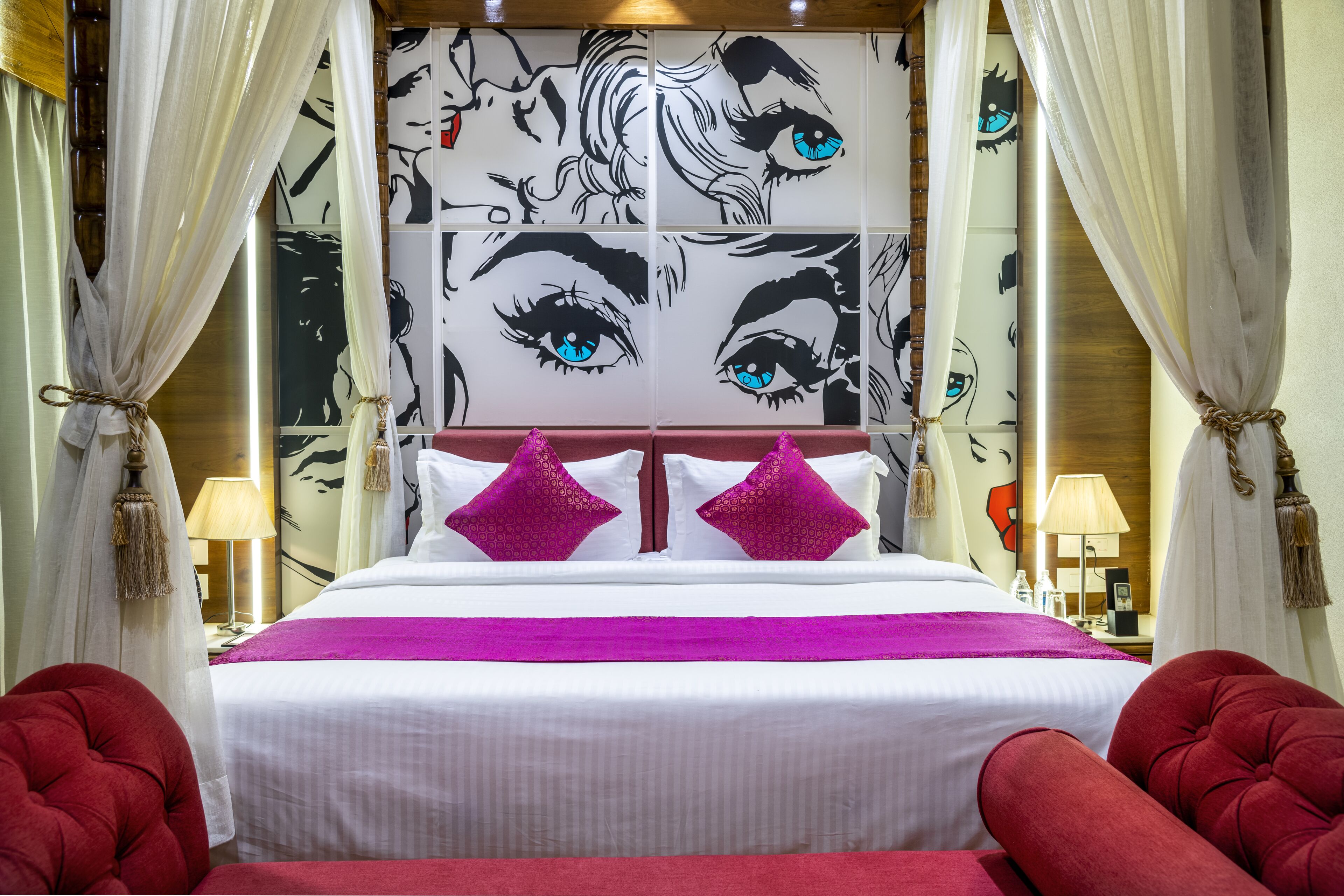 Ein Bett mit Kopfteil, das ein großes, abstraktes Gesichtsmotiv zeigt, zwei rote Sessel und eine pinkfarbene Decke.