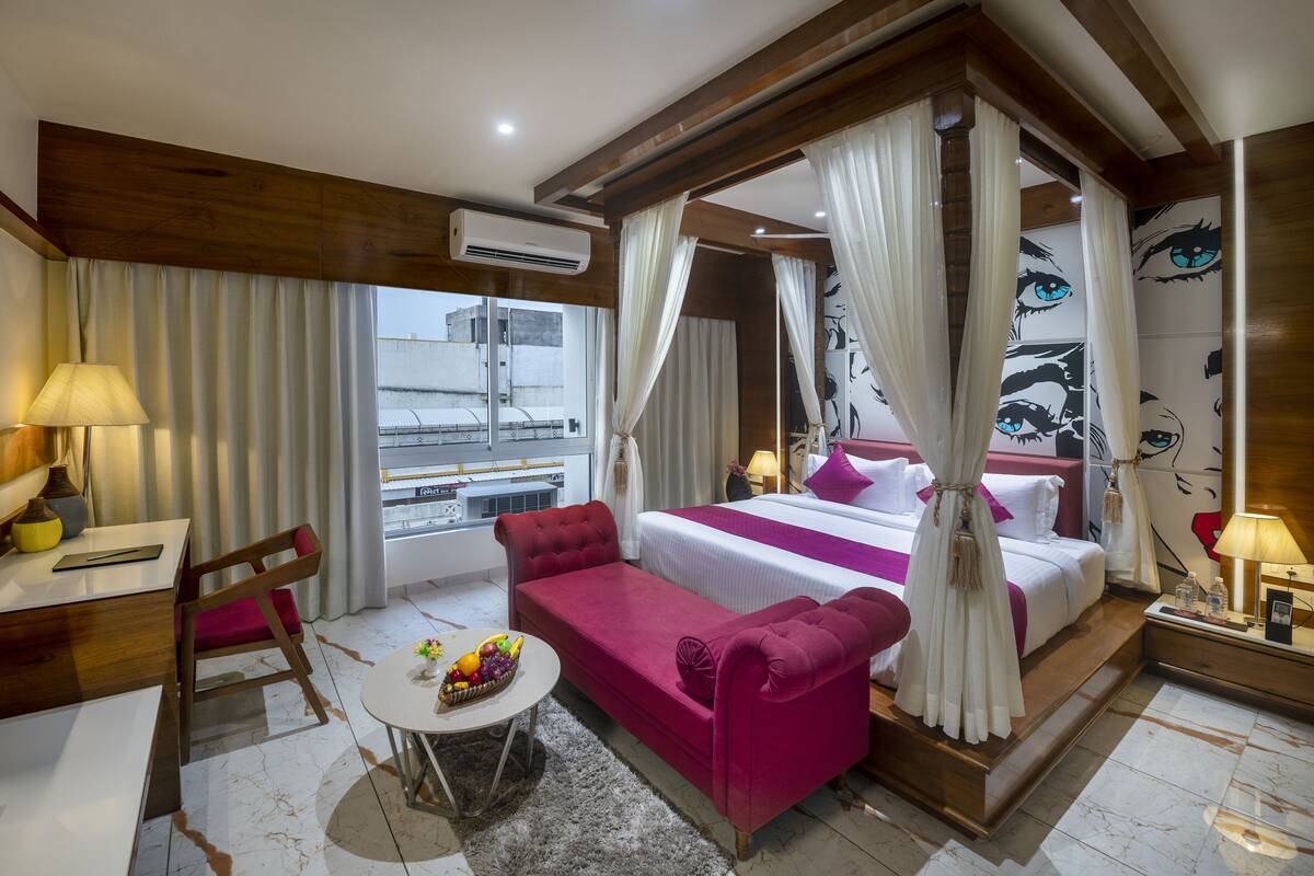 A hotel room with a bed, a desk, a chair, a small table with fruit, and a view of the outside.