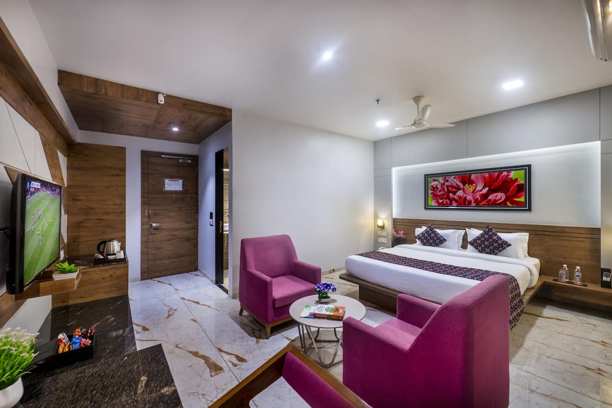 A modern hotel room with a large bed, two pink armchairs, a small table, a TV, and a floral artwork on the wall.