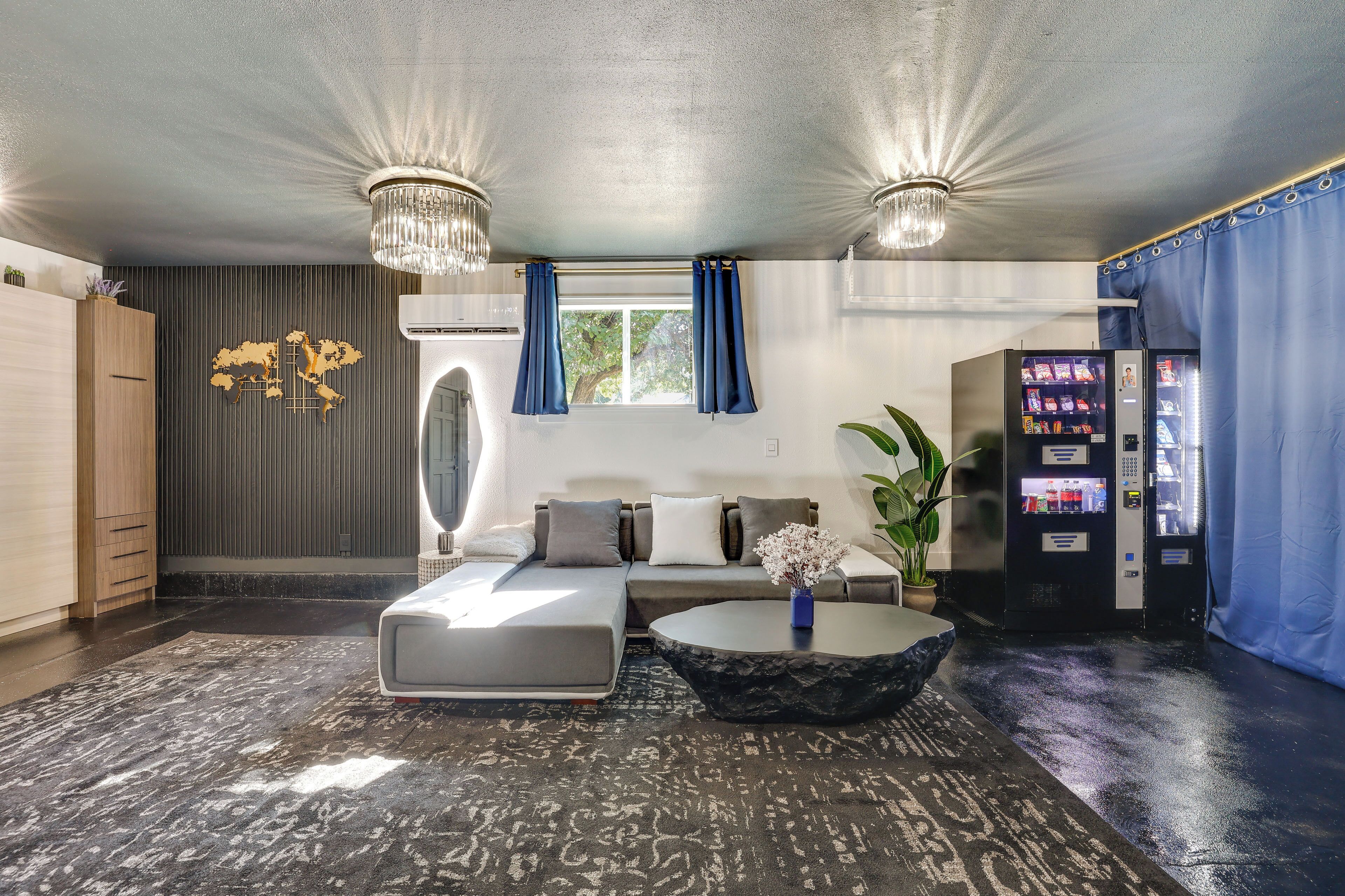 Ein modernes Hotelzimmer mit Sofa, Couchtisch, Wanddeko, einem Fenster mit Vorhängen und einer Getränkeautomaten.