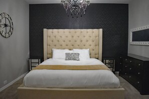 A bedroom with a tufted headboard, a large bed, a chandelier, a clock, and a dresser.