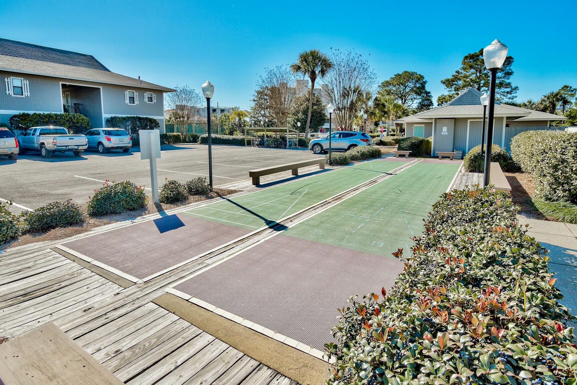 A parking lot with several cars, a green sports court, a wooden deck, and a small building with a covered area.