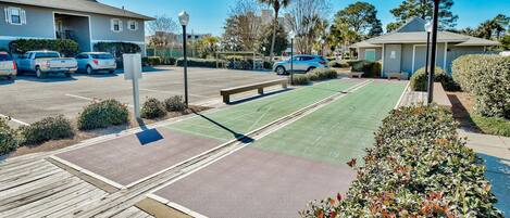 A parking lot with several cars, a green sports court, a wooden deck, and a small building with a covered area.