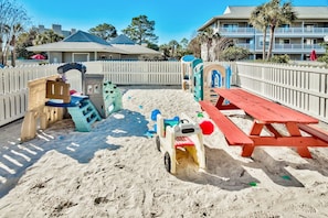A sandy play area with a wooden bench and a picnic table, surrounded by a white fence.