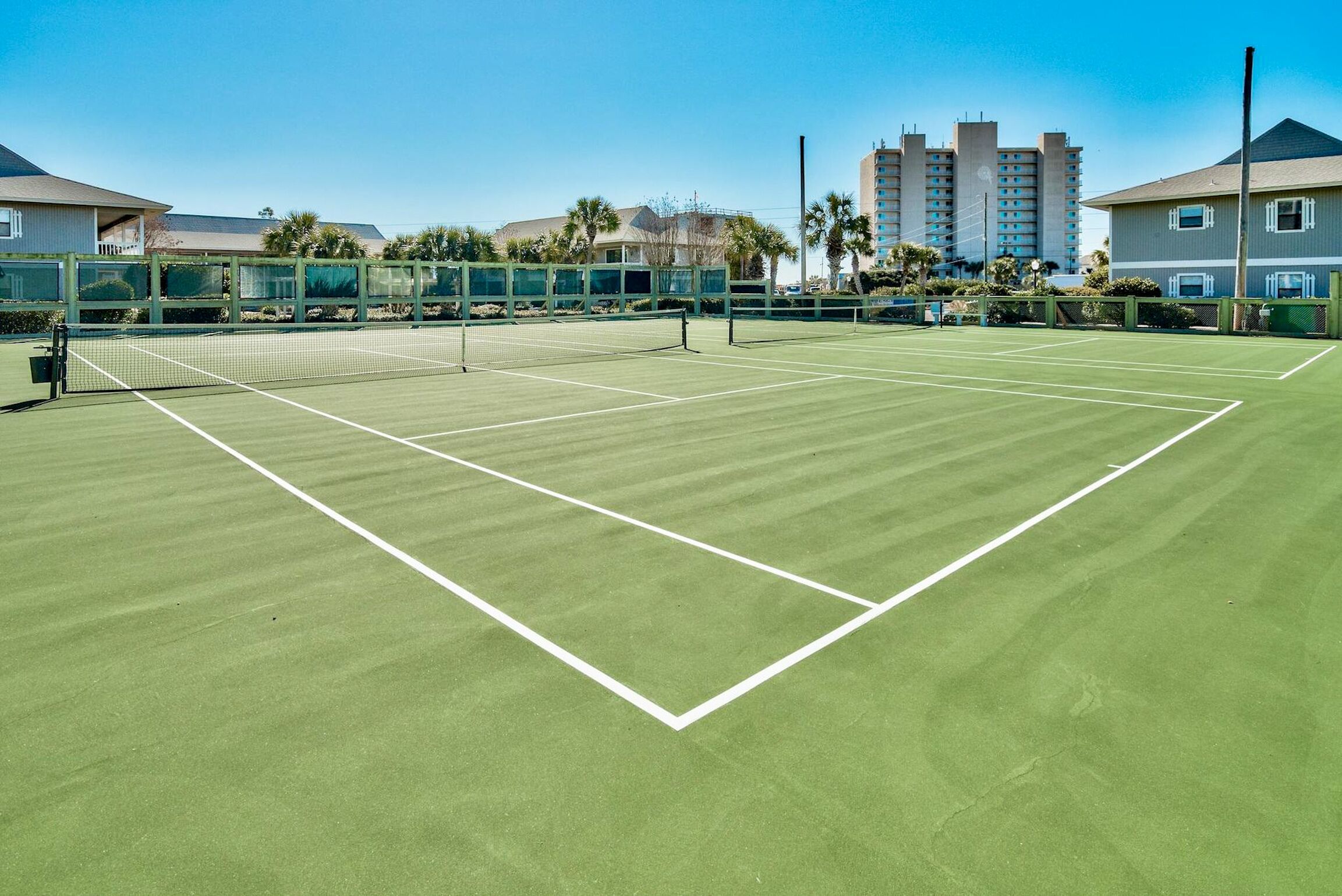 Un court de tennis avec un filet au milieu, entouré d’une clôture, et des bâtiments en arrière-plan.