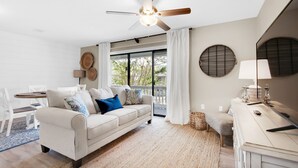 Ein modernes Wohnzimmer mit einem weißen Sofa, einem Deckenventilator, einem Esstisch und einem großen Fenster mit Vorhängen.