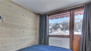A bedroom with a wooden wall, a bed with a blue bedspread, a window with a view of snow-covered mountains, and a radiator.