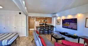 A living room with a sofa, a bench, a dining table with chairs, a kitchen area, and a mounted TV. - Entire Condominium - Newly upgraded Waikiki TOP Floor ILIKAI Hotel (Honolulu)