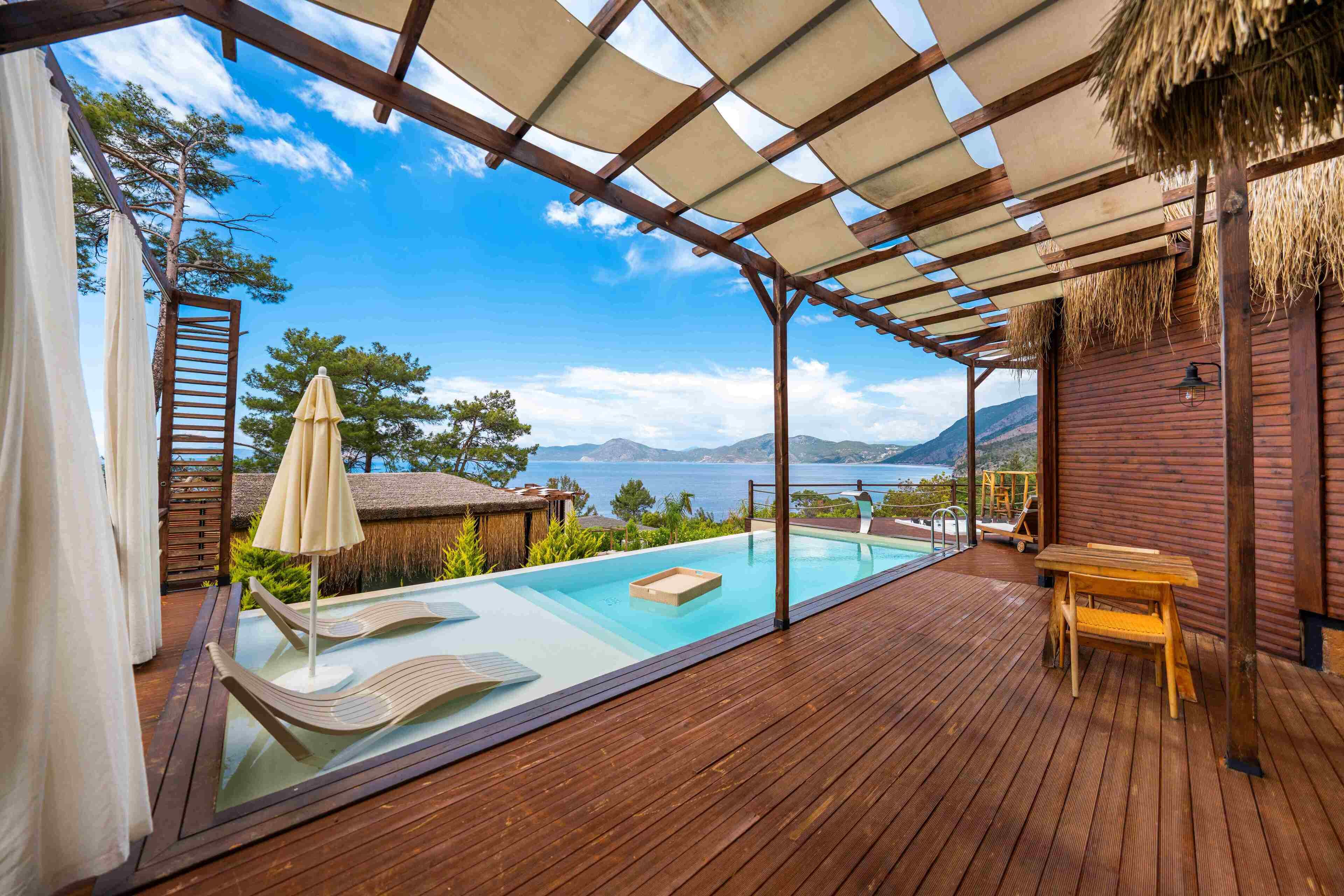 A wooden deck with a glass-bottomed infinity pool, a wooden table, and a lounge chair.