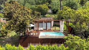 Un jacuzzi rodeado por una terraza de madera, asientos exteriores y abundante vegetación.