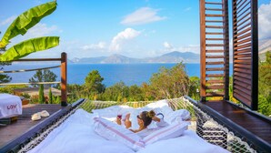 A hammock bed with a view of the sea and mountains, a wooden deck, and a 'LOV' towel.