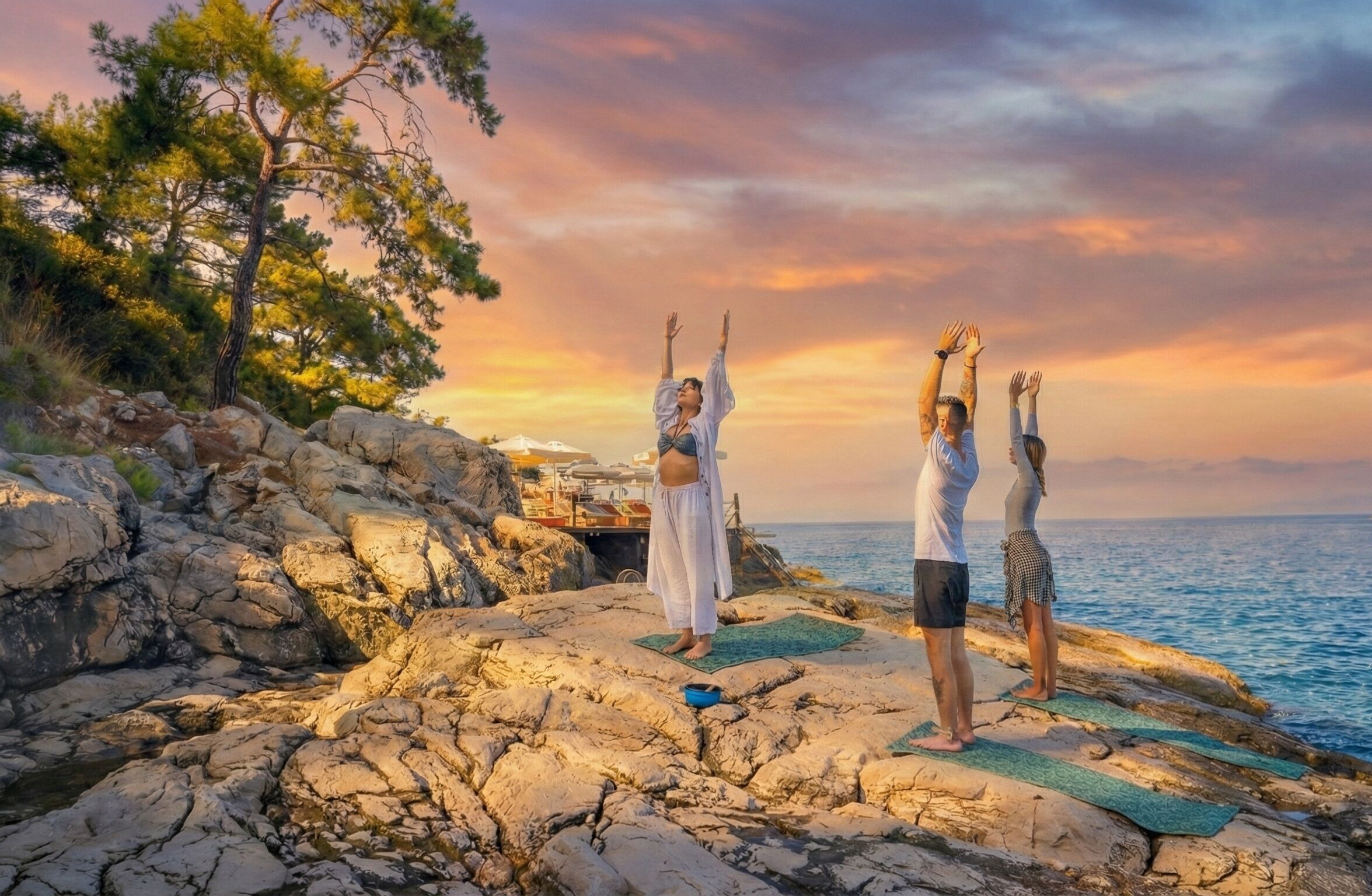 Tres personas practicando yoga sobre terreno rocoso, con vista al mar y al atardecer.