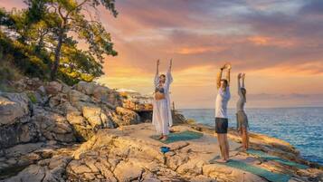 Tres personas practicando yoga sobre terreno rocoso, con vista al mar y al atardecer.