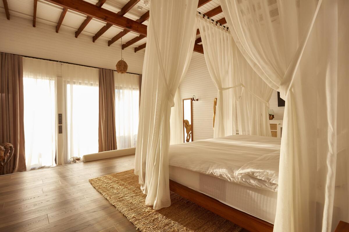 Une chambre à coucher avec un lit à baldaquin, des poutres apparentes, une grande fenêtre avec des rideaux et un tapis tissé.