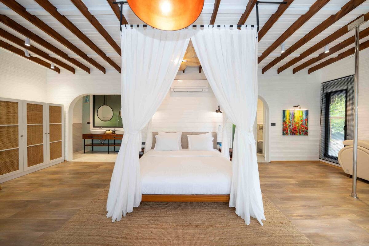 Une chambre à coucher avec un lit à baldaquin, des poutres en bois, une grande fenêtre et une salle de bain équipée d’un lavabo et d’un miroir.