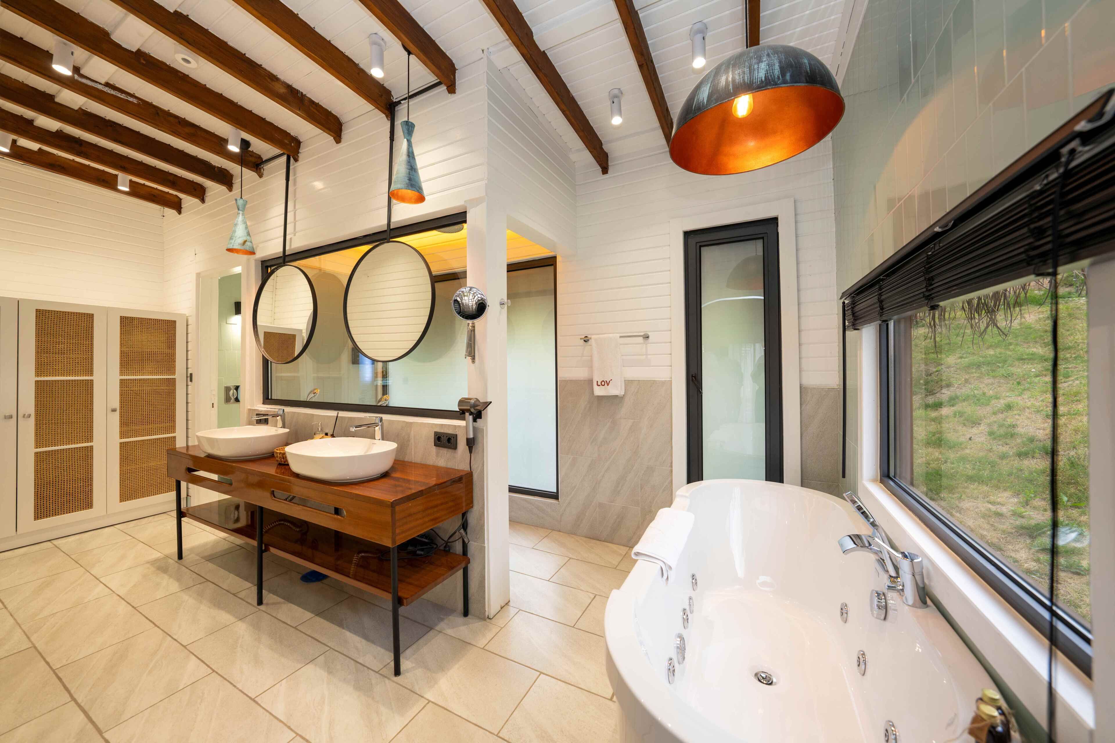 Une salle de bain moderne avec deux vasques, une grande baignoire, des poutres en bois et une vue par la fenêtre.
