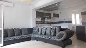 A modern living room with a grey sectional sofa, a kitchen area with dark cabinets, and a large window providing natural light. - S+2 close to the sea chott meriem (Sousse)