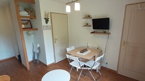 Una habitación pequeña y bien iluminada con piso de madera, una mesa blanca con cuatro sillas, una mesita blanca, un televisor montado en la pared y una puerta con un letrero encima.
