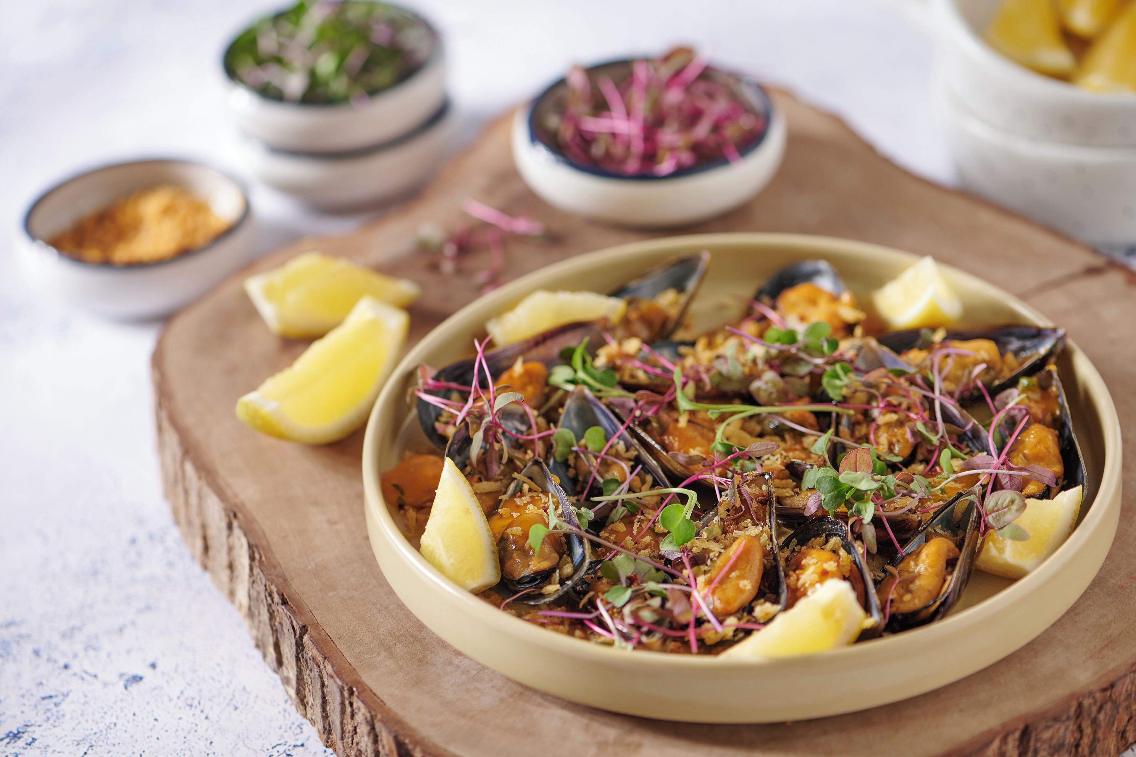 A bowl of mussels garnished with lemon and microgreens, with additional bowls of condiments and lemon wedges in the background.