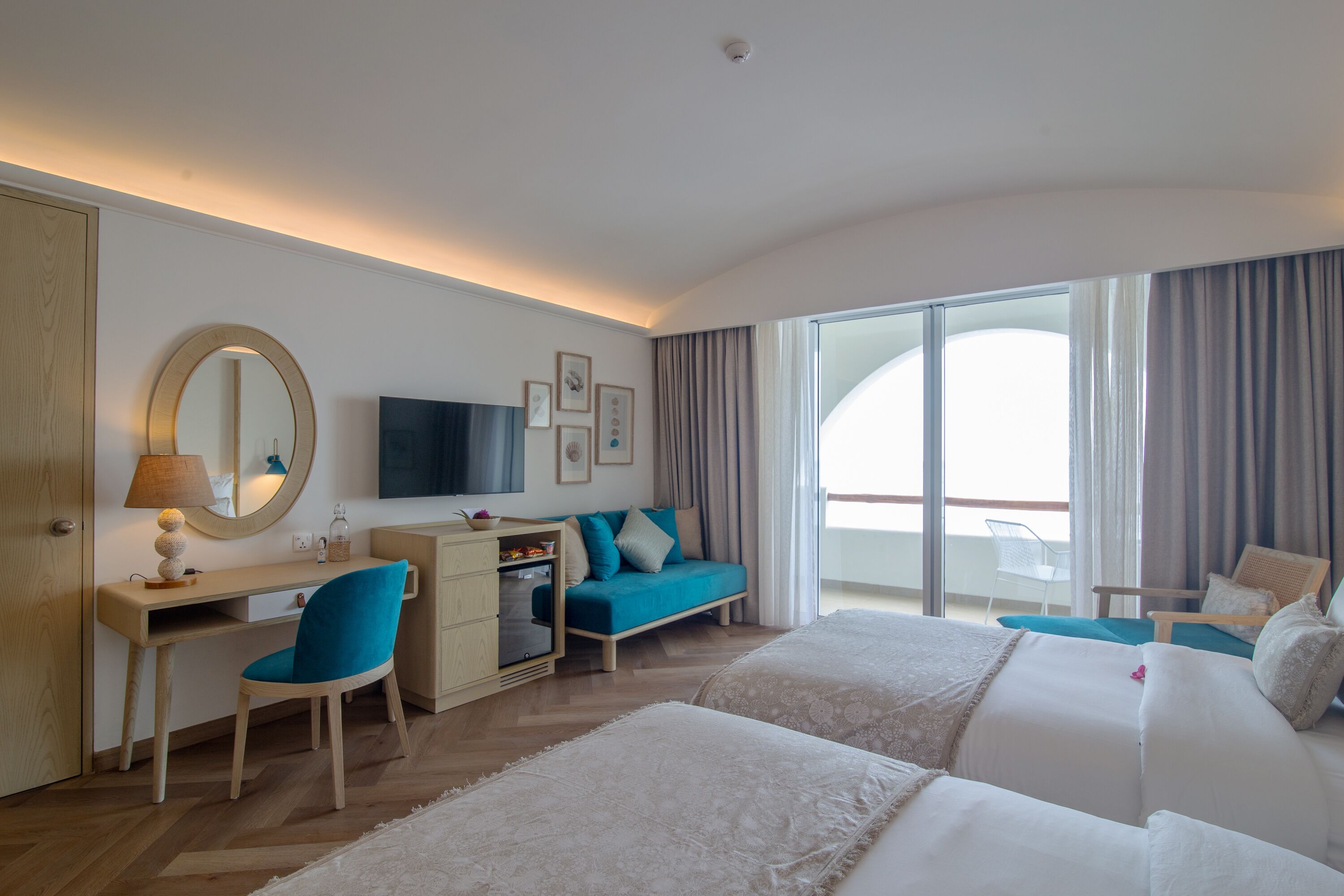 Habitación de hotel con cama, escritorio con silla, televisor, sofá y balcón con cortinas.