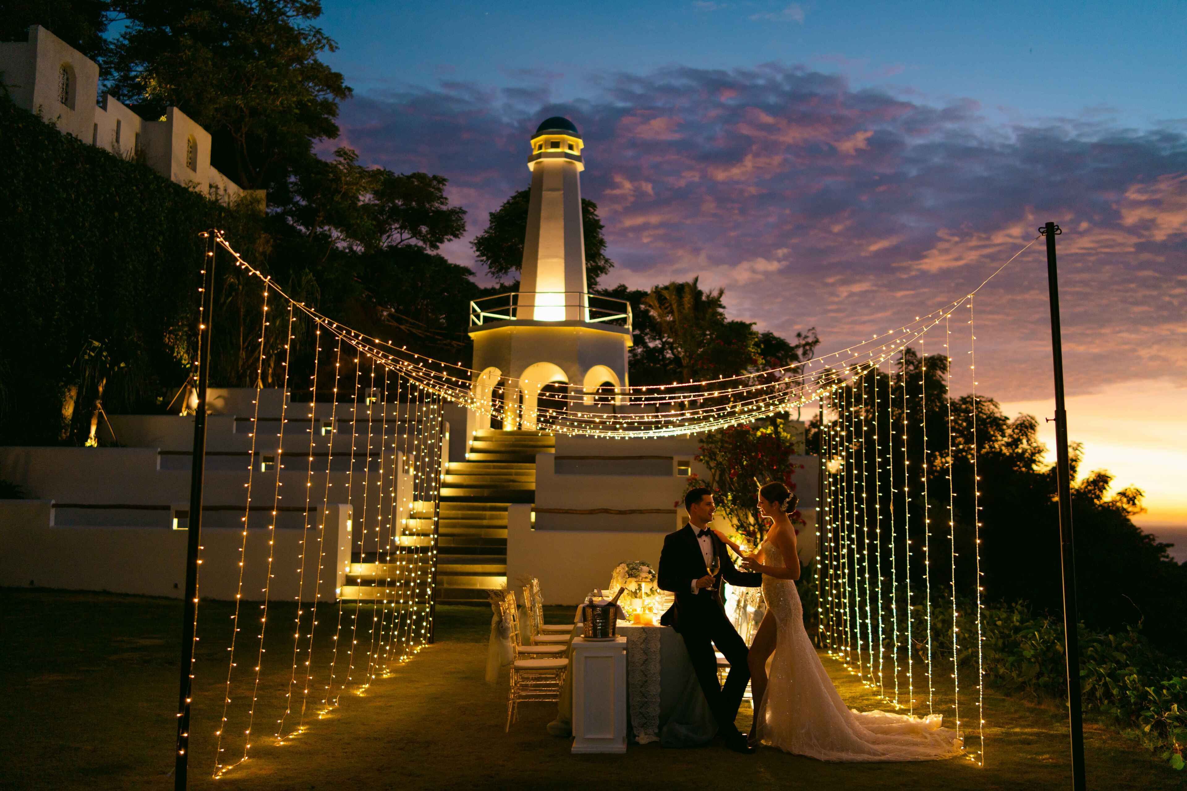 A wedding ceremony with a couple seated at a table, surrounded by string lights and a lighthouse in the background.