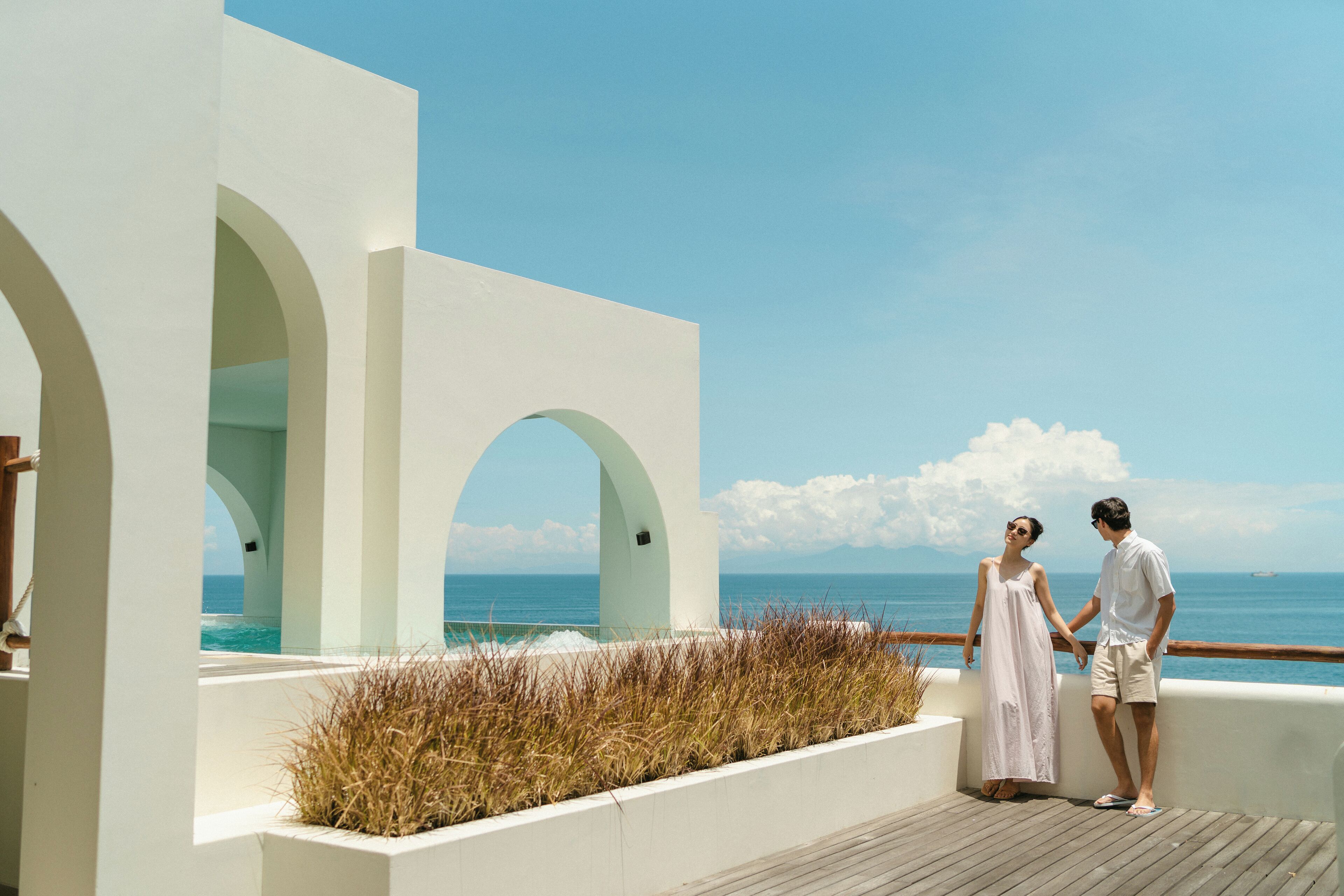 A couple standing on a wooden deck overlooking the ocean, with white architecture and arches.