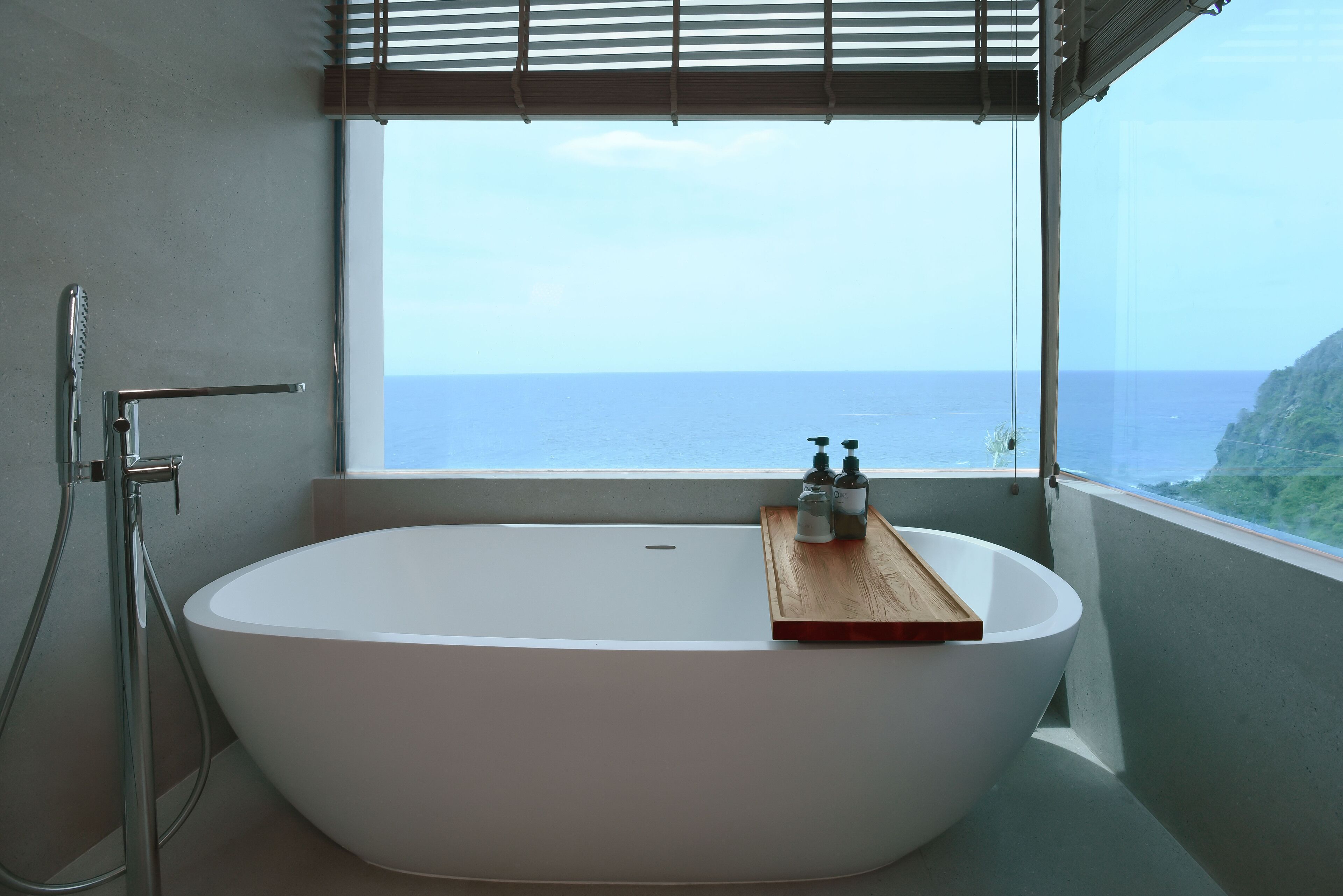 A modern bathroom with a freestanding bathtub, a shower, and a window offering a view of the sea.
