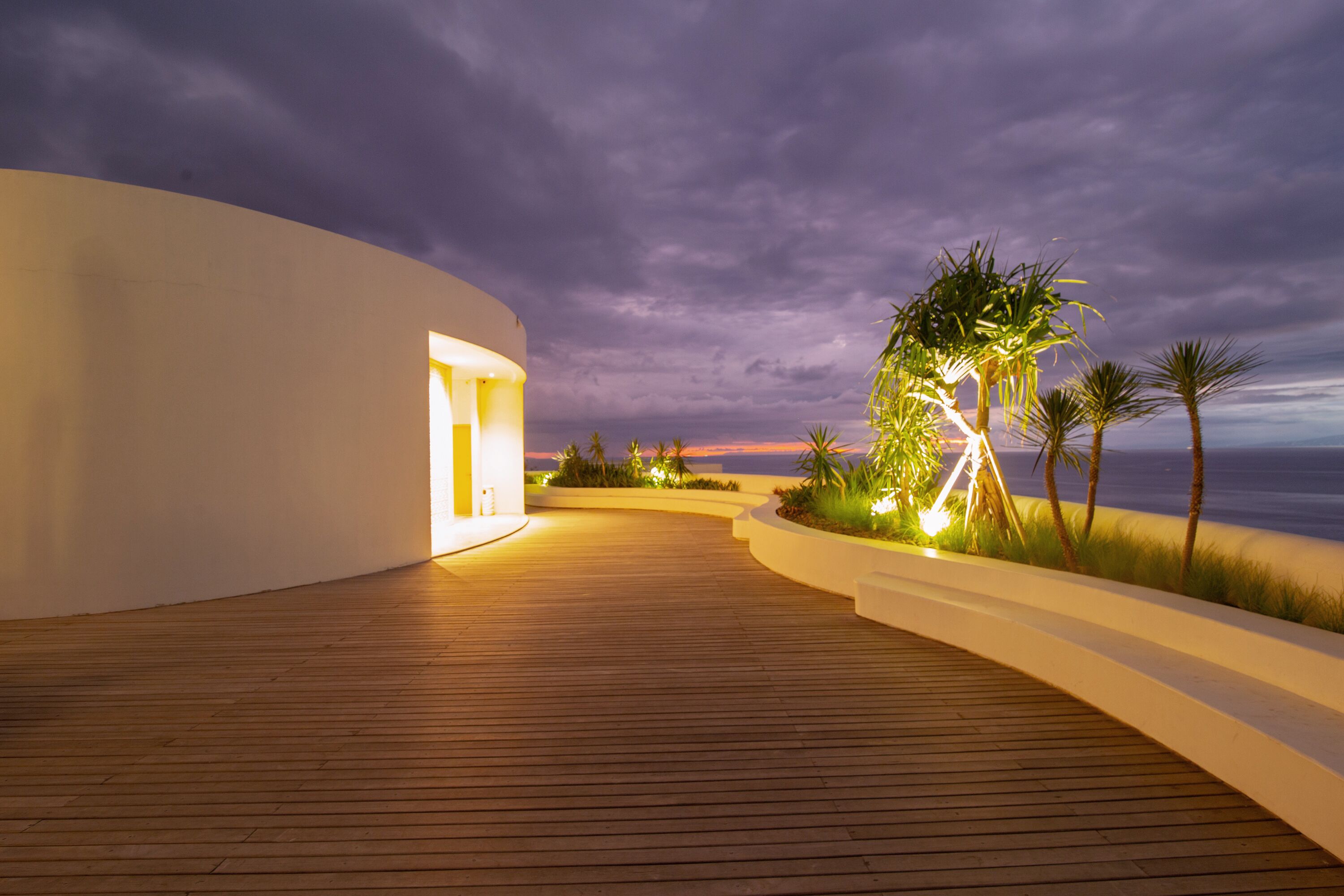 A wooden deck leading to a white building with a curved facade, surrounded by palm trees and a view of the ocean at dusk.