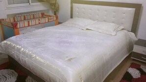 Cama bem arrumada com edredom branco e travesseiros, mesa de cabeceira e estante com livros.