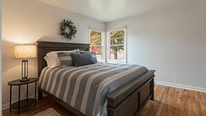 A bedroom with a bed, a nightstand, a lamp, a wooden floor, a window, and a door. - Cute Home in the Heart of Missoula! (Missoula)