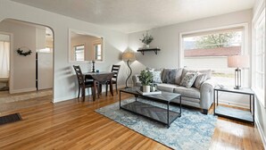 A living room with a sofa, coffee table, and dining table. There is a window with a view of the outside. - Cute Home in the Heart of Missoula! (Missoula)