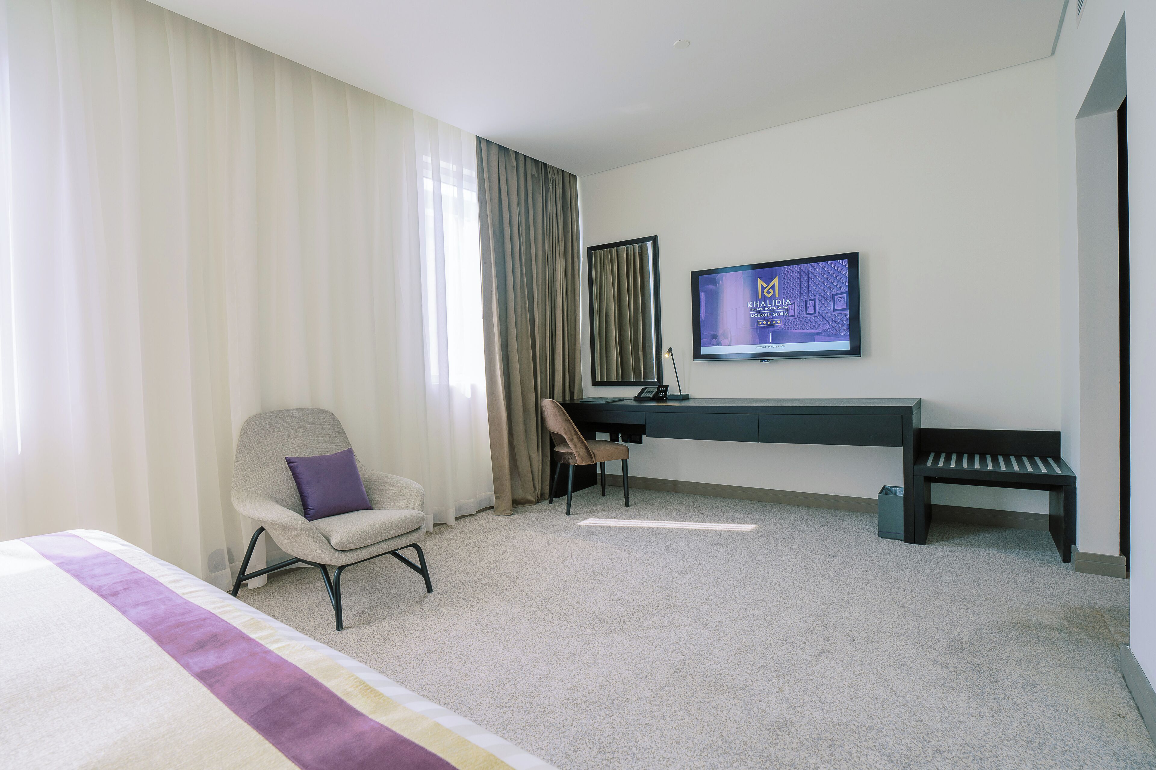 A hotel room with a bed, a desk, a chair, a television, and a bench.