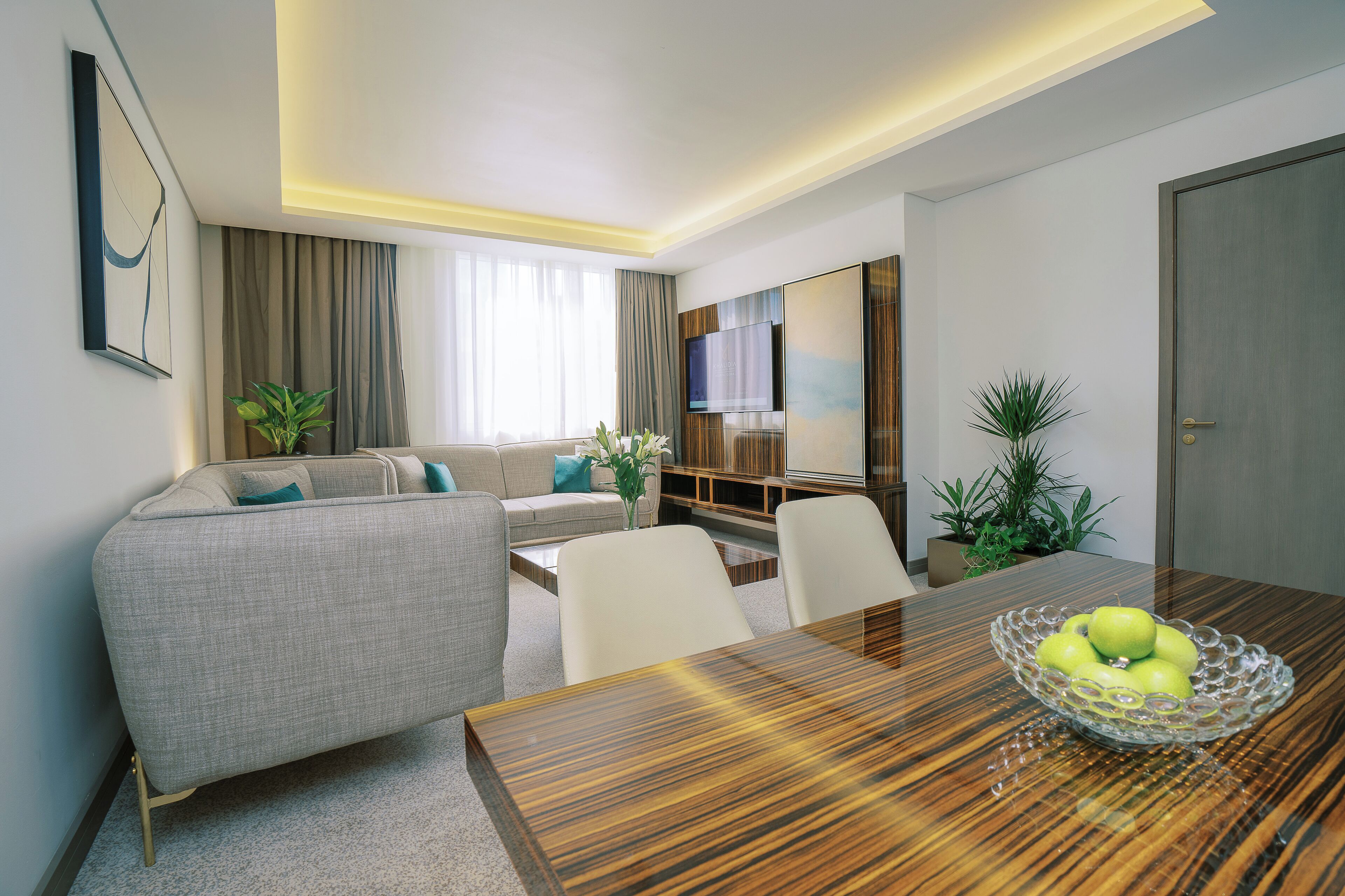A modern hotel room with a wooden dining table, a sofa, a TV, and a plant.