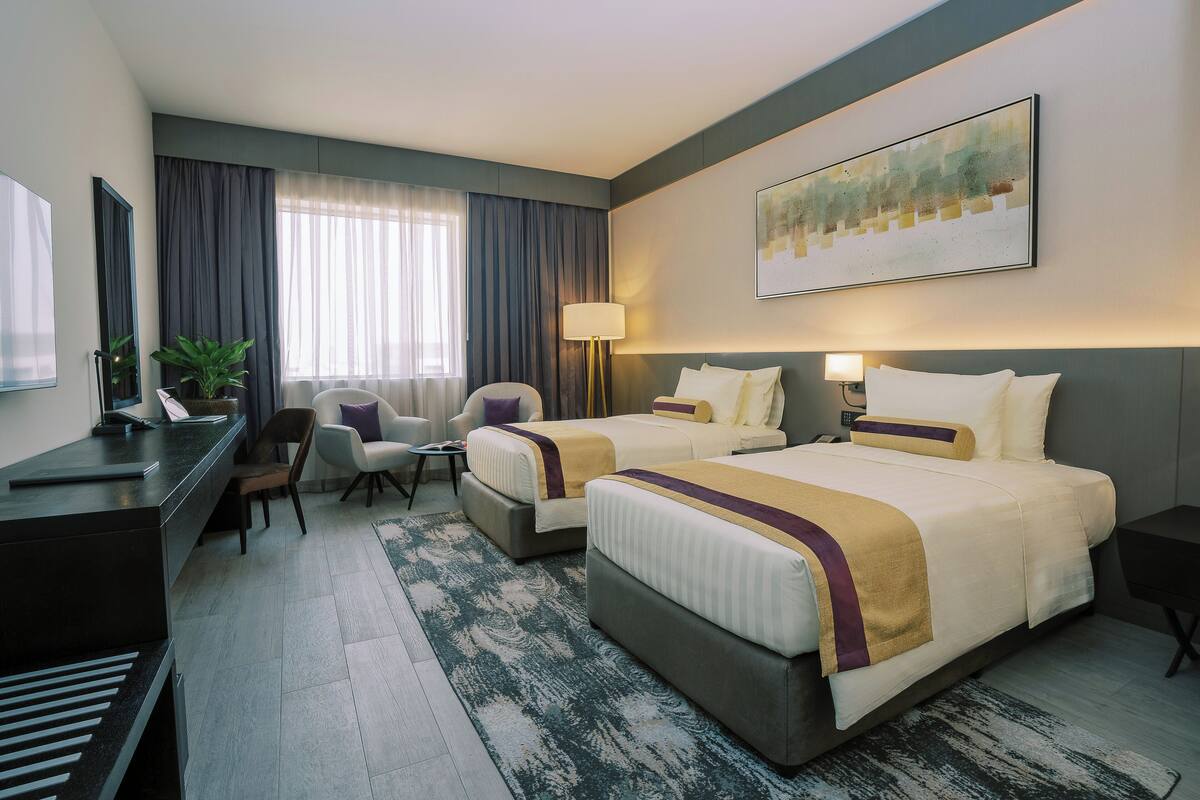 A hotel room with two beds, a desk, a chair, a lamp, and a painting on the wall.
