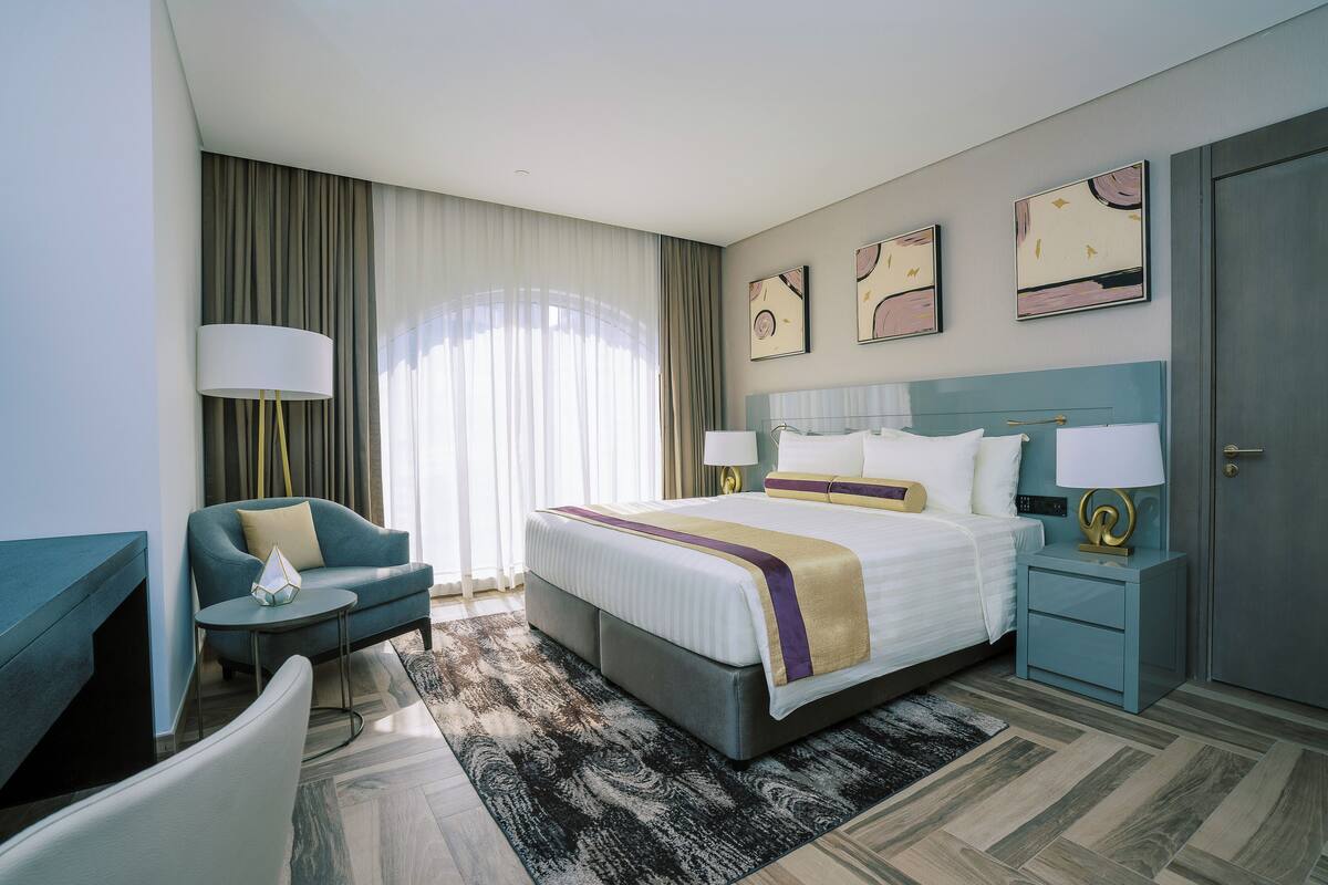 A modern hotel room with a large bed, a seating area with a chair and a small table, a nightstand with a lamp, and artwork on the walls.