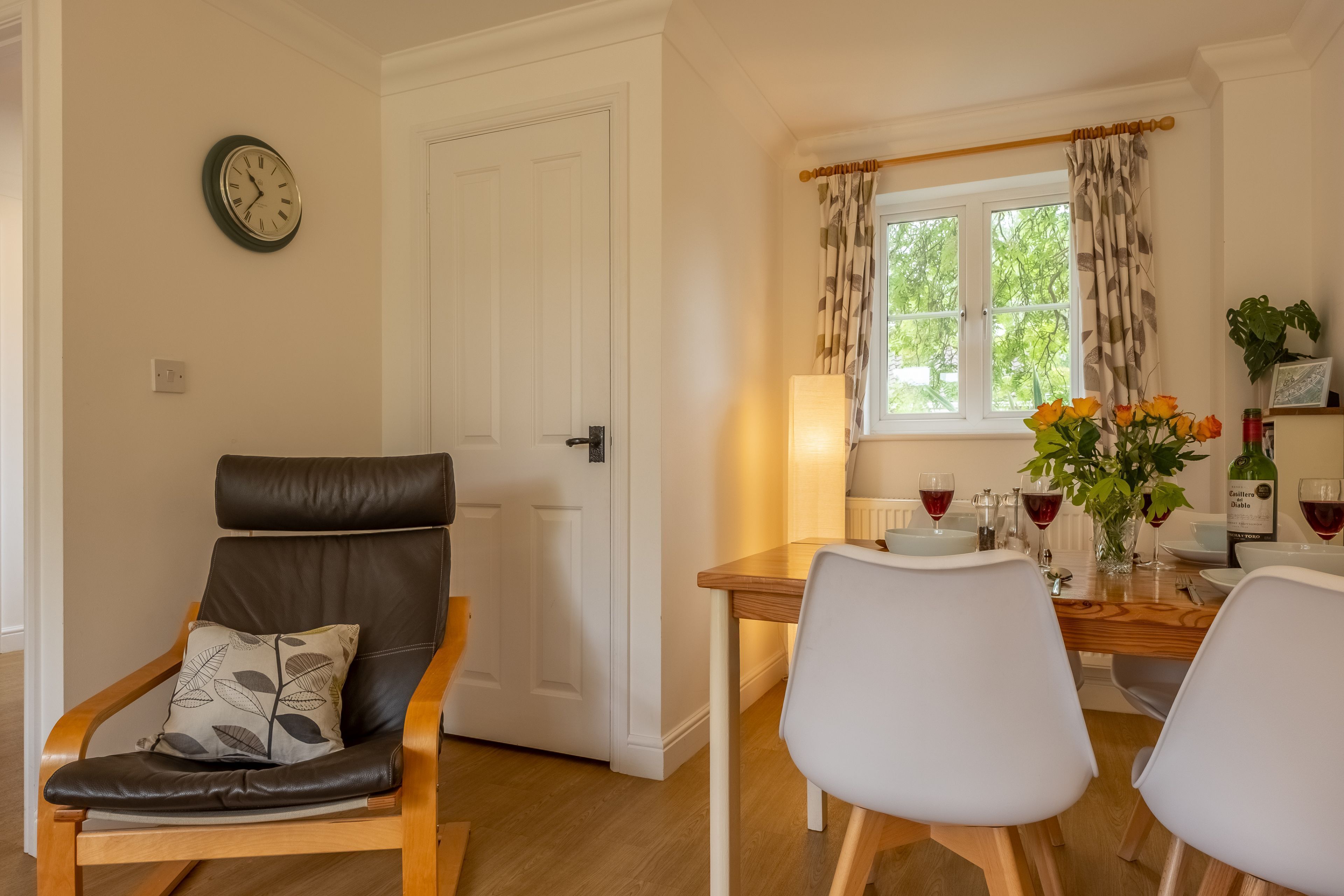 A dining area with a wooden table, two chairs, a clock on the wall, and a window with curtains.