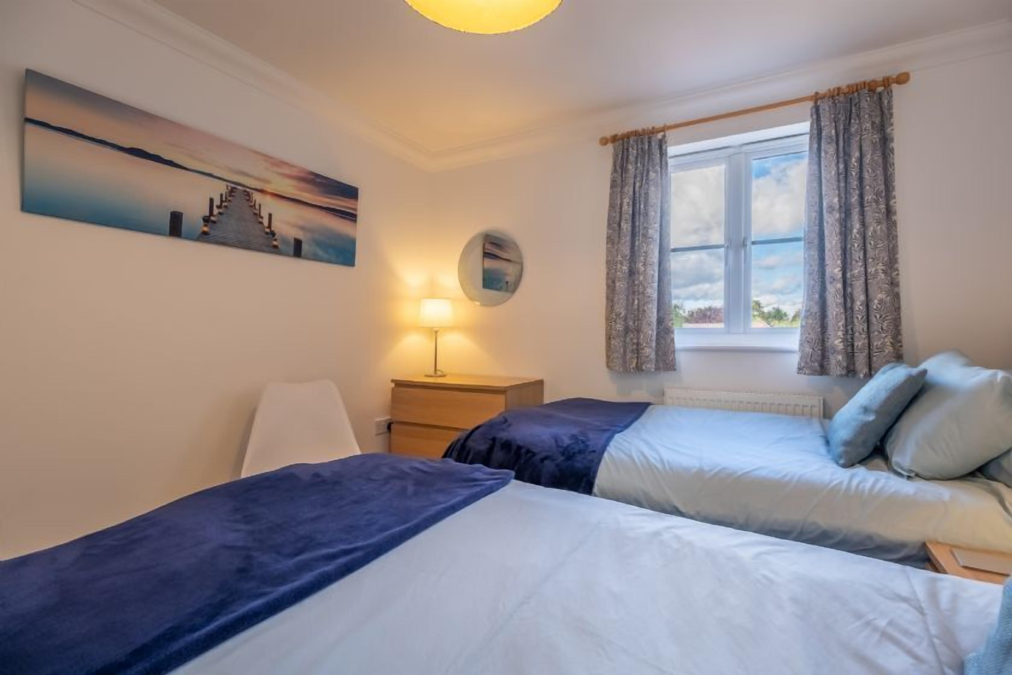 A bedroom with two beds, a nightstand, a lamp, a window with curtains, and a painting on the wall.