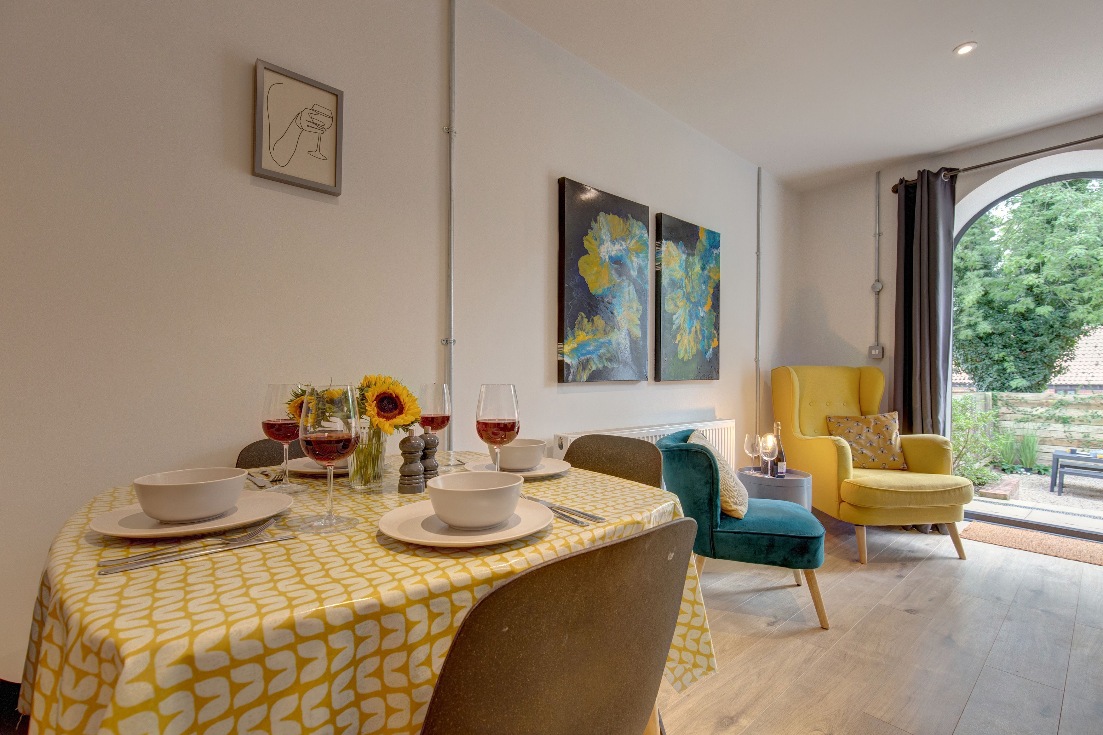 Un coin repas avec une table dressée pour deux personnes, un fauteuil jaune et une vue sur la verdure à l’extérieur.