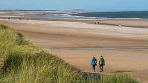 Ein weiter Sandstrand mit Menschen, die gehen und stehen, und eine Rasenfläche im Vordergrund.