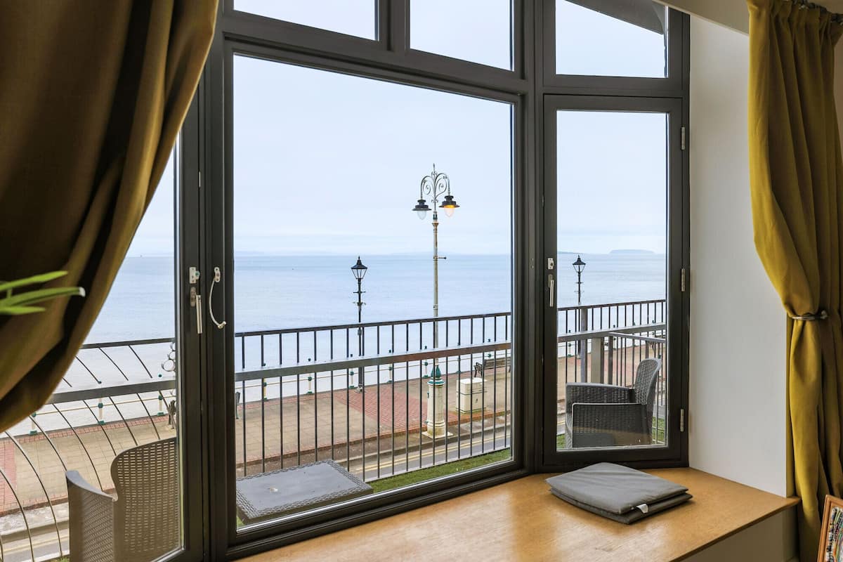 A room with a large window overlooking a seaside view, a small table with a cushion, and a metal railing.