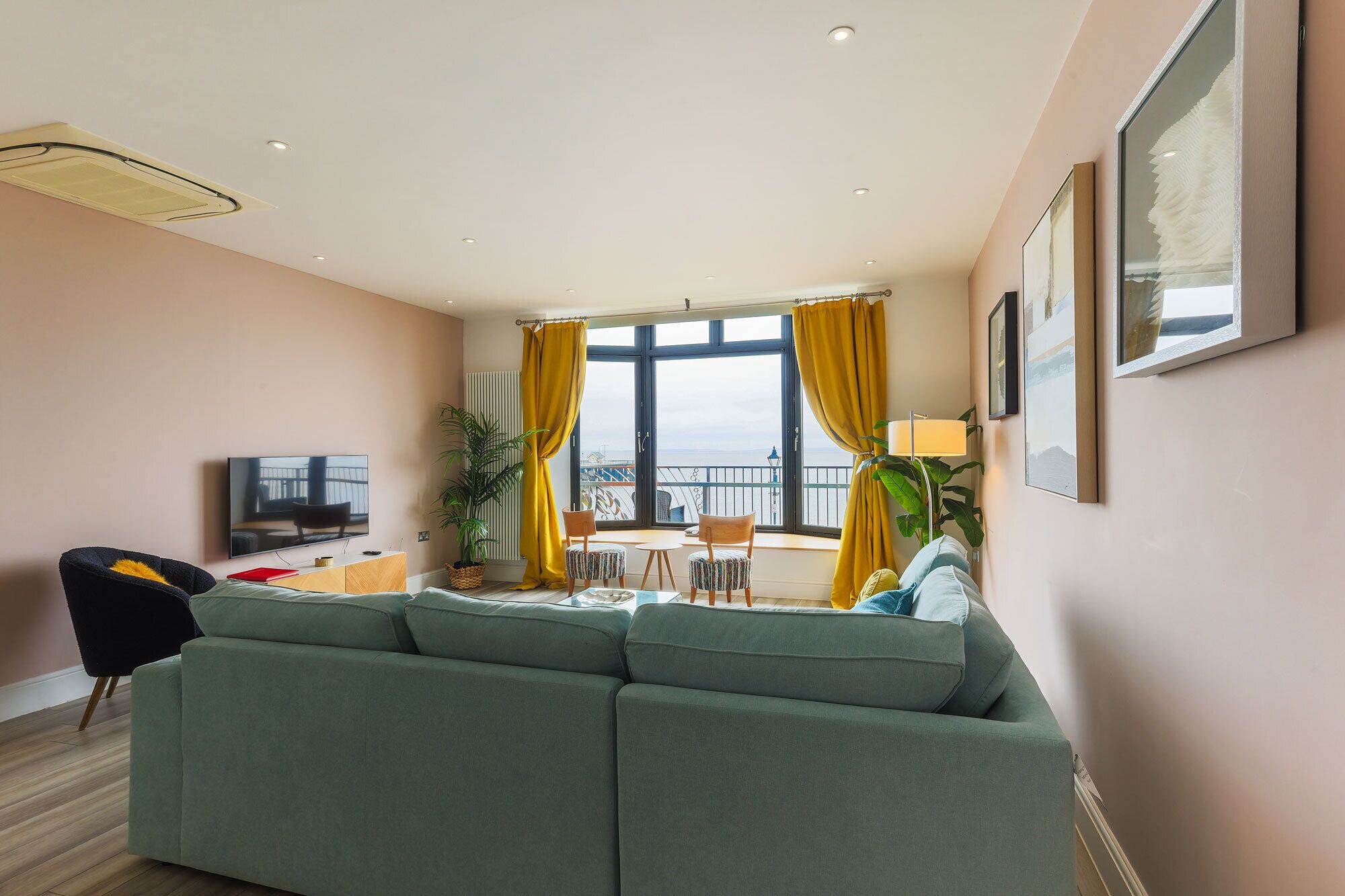 Un salon avec un canapé, un téléviseur, un balcon avec des rideaux jaunes et une plante.