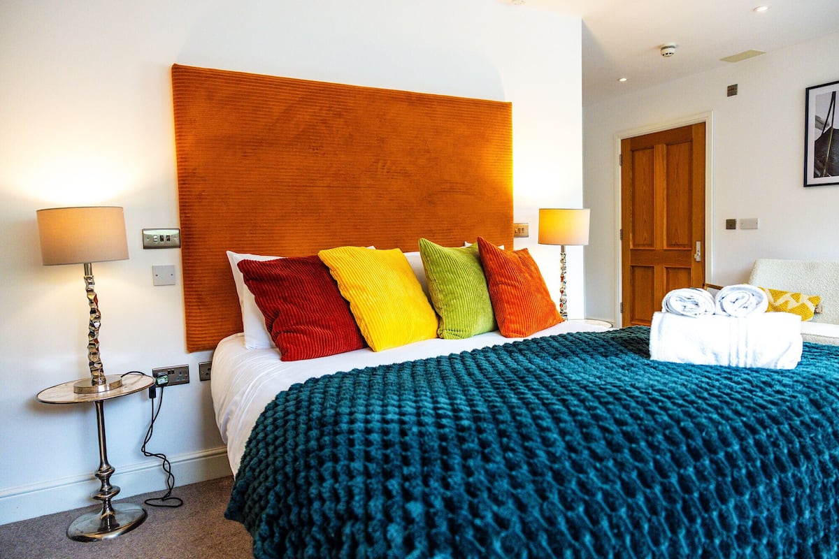 A hotel room with a large bed, colorful pillows, a blue blanket, and two bedside lamps.