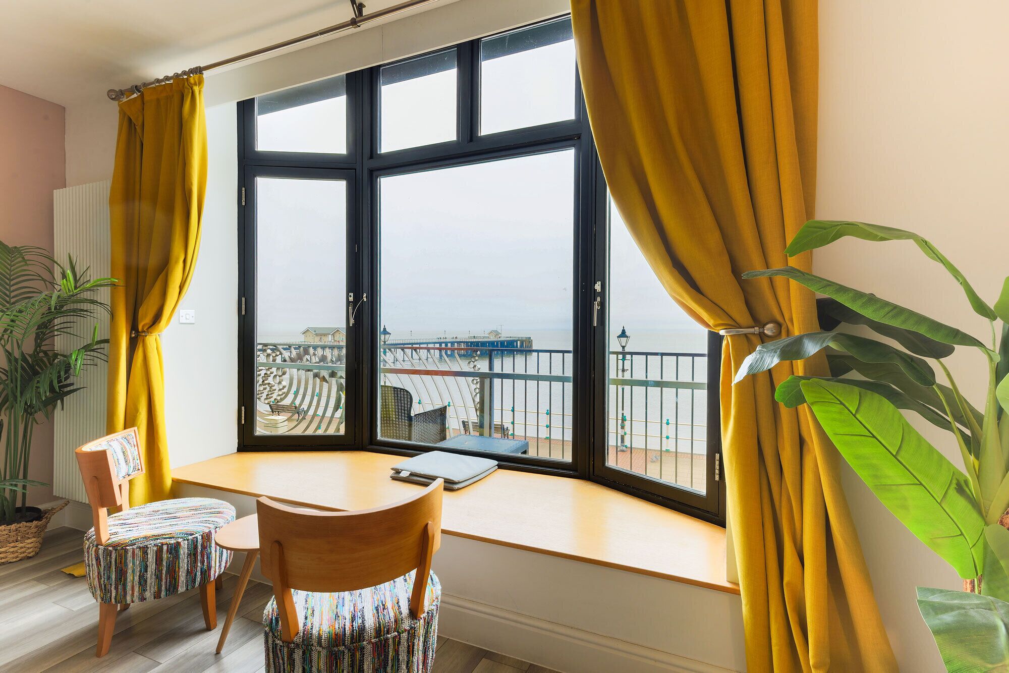 A room with a large window, a wooden table, two chairs, and a view of a pier.