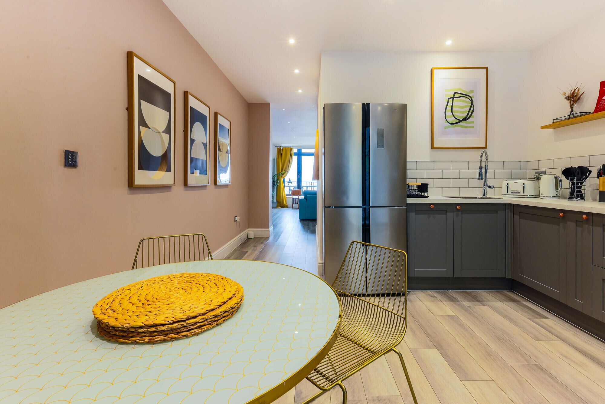 A modern kitchen with a stainless steel refrigerator, a dining area with a round table and chairs, and a hallway leading to a living space.